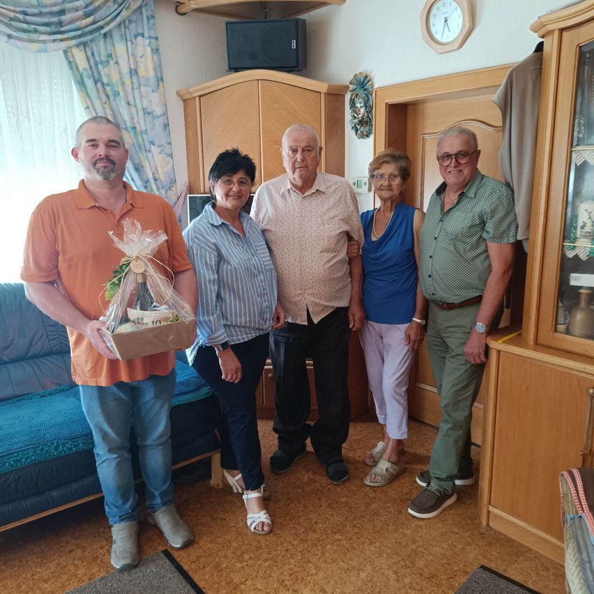 Fünf Menschen stehen lächelnd für ein Foto im Wohnzimmer. Ein Mann hält einen Geschenkkorb. Hinter ihnen sind eine Uhr und ein Regal mit Gegenständen zu sehen.