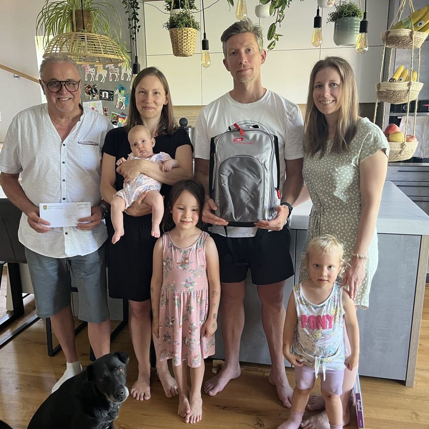 Eine Familie mit fünf Personen posiert für ein Foto in einer Küche mit einem Hund, Bananen und Pflanzen. Ein Mann hält einen Rucksack.
