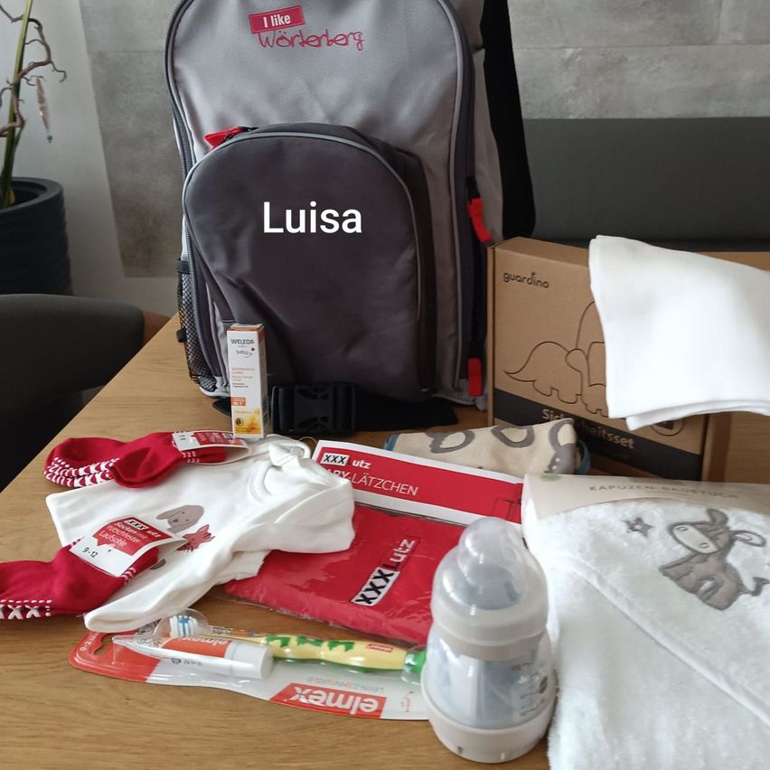 Auf einem Tisch stehen ein Babyrucksack namens Luisa, eine Schachtel, ein Handtuch, Babykleidung, eine Zahnbürste und eine Flasche.