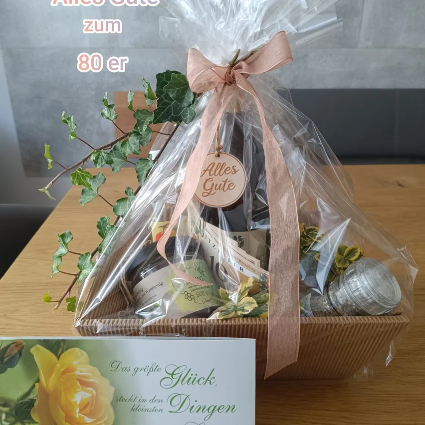 Ein Geschenkkorb mit einem rosa Band und einem Schild 'Alles Gute' ist in Plastik verpackt und enthält verschiedene Artikel wie eine Flasche, ein Glas und eine kleine Pflanze.