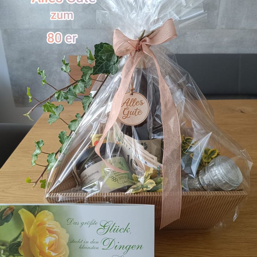 Ein Geschenkkorb mit einem rosa Band und einem Schild 'Alles Gute' ist in Plastik verpackt und enthält verschiedene Artikel wie eine Flasche, ein Glas und eine kleine Pflanze.