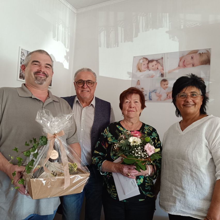 Vier Menschen stehen lächelnd in einem Raum. Ein Mann hält eine Geschenkbox mit einem Band, und eine Frau hält einen Blumenstrauß. Hinter ihnen zeigt eine Wand Familienfotos.
