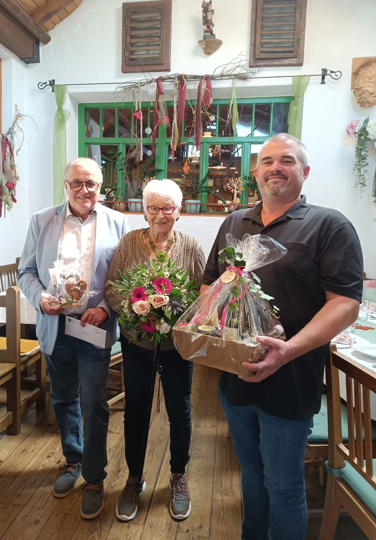 Drei Personen stehen zusammen in einem Raum, jeder hält ein Geschenk. Der Mann auf der linken Seite trägt eine Brille und eine hellblaue Jacke, die Frau in der Mitte hält einen Blumenstrauß und der Mann auf der rechten Seite hält eine Schachtel mit einem Band.