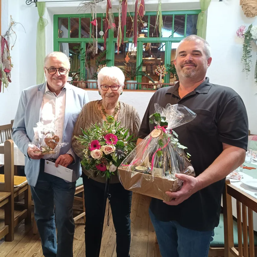 Drei Personen stehen zusammen in einem Raum, jeder hält ein Geschenk. Der Mann auf der linken Seite trägt eine Brille und eine hellblaue Jacke, die Frau in der Mitte hält einen Blumenstrauß und der Mann auf der rechten Seite hält eine Schachtel mit einem Band.