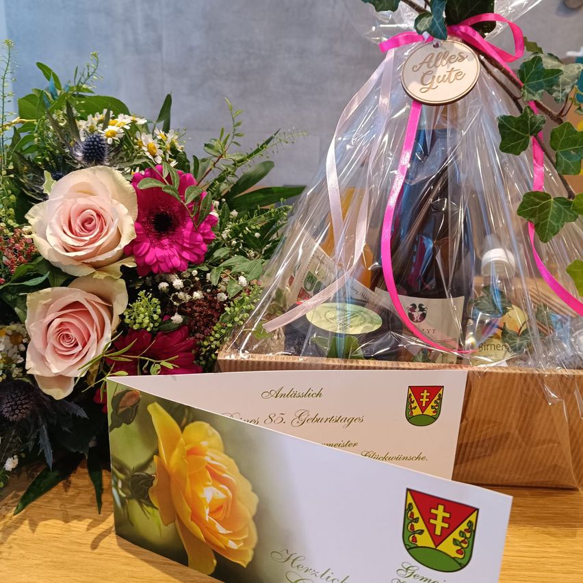 Ein Blumenstrauß und eine Flasche Wein in einer durchsichtigen Plastiktüte mit einem rosa Band, zusammen mit einer Karte, auf der 'Herzlichen Gluckwunsch' steht, auf einem Holztisch.