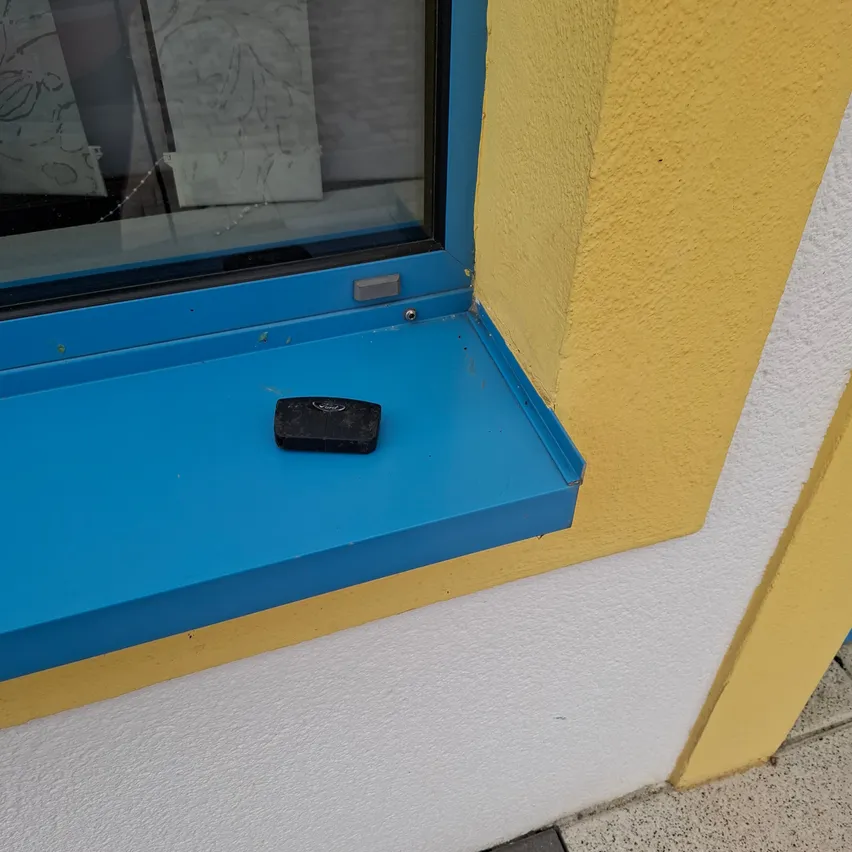 Ein schwarzes Objekt, möglicherweise eine Fernbedienung, befindet sich auf der blauen Fensterbank eines Gebäudes.