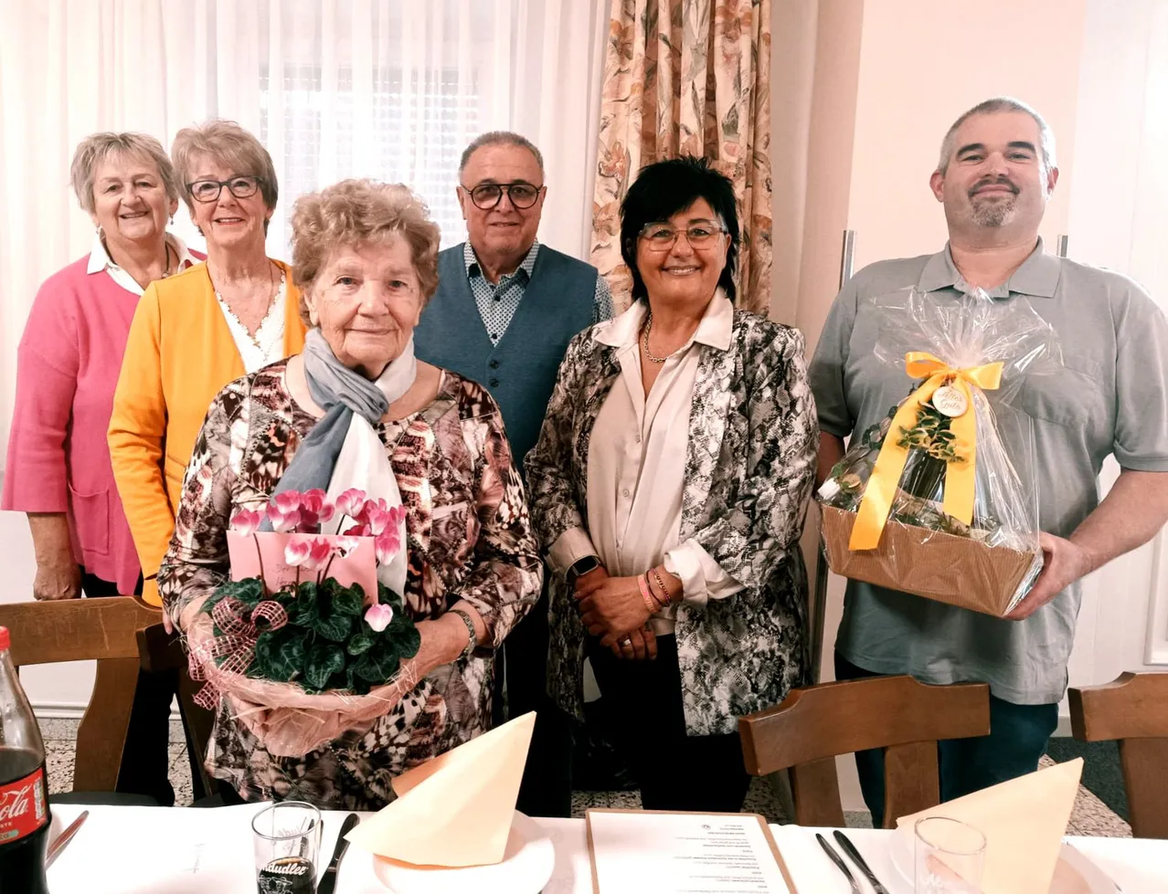 Eine Gruppe von Menschen steht hinter einem Tisch und lächelt für ein Foto. Eine Frau in der Mitte hält einen Blumenstrauß.
