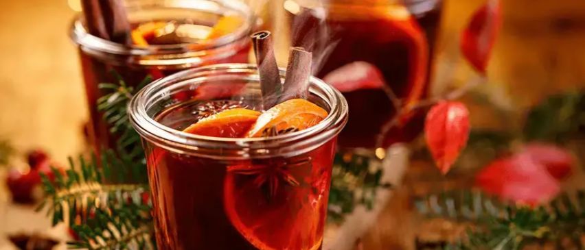 Drei Gläser Glühwein mit Orangen und Zimt, die Dampf abgeben. Die Umgebung ist festlich mit roten Blättern und Beeren auf einer Holzoberfläche.