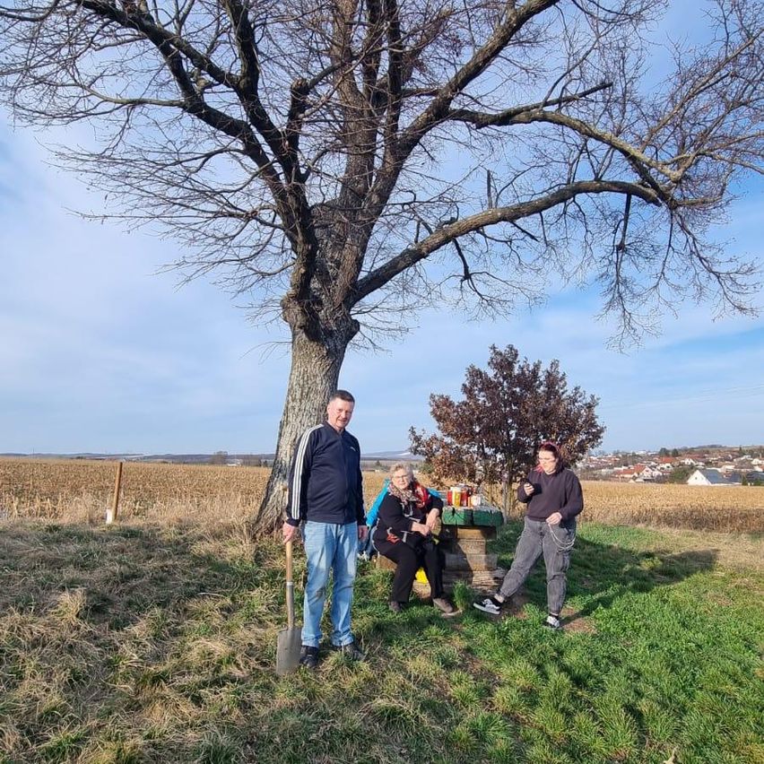 Drei Personen stehen draußen mit einer Bank und einem Baum im Hintergrund. Der Mann hält eine Schaufel.