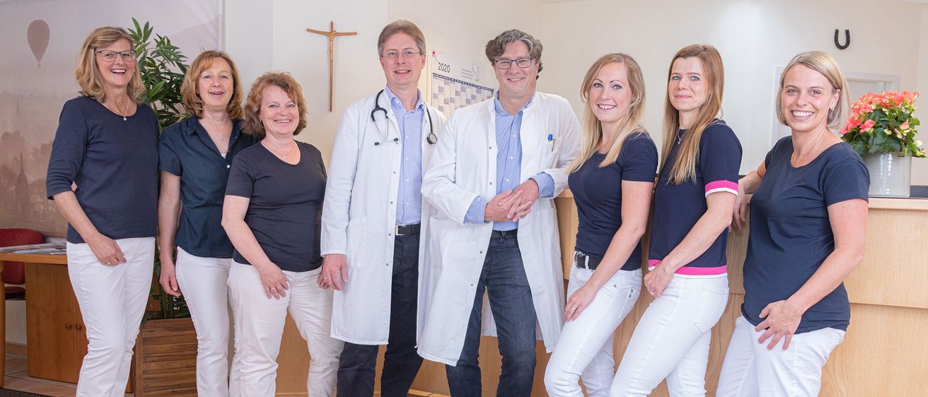 Eine Gruppe von medizinischen Fachkräften steht gemeinsam in einem Raum. Sie tragen alle blaue Uniformen und lächeln. Die Männer sind in weißen Laborkittel. Hinter ihnen befindet sich eine Wand mit einem Kalender und einem Kreuz.