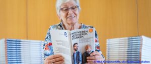 Eine ältere Frau lächelt und liest ein Buch mit dem Titel Motivieren Aktivieren Starken. Auf dem Cover des Buches sind zwei Männer abgebildet. Dahinter befinden sich Stapel desselben Buches.