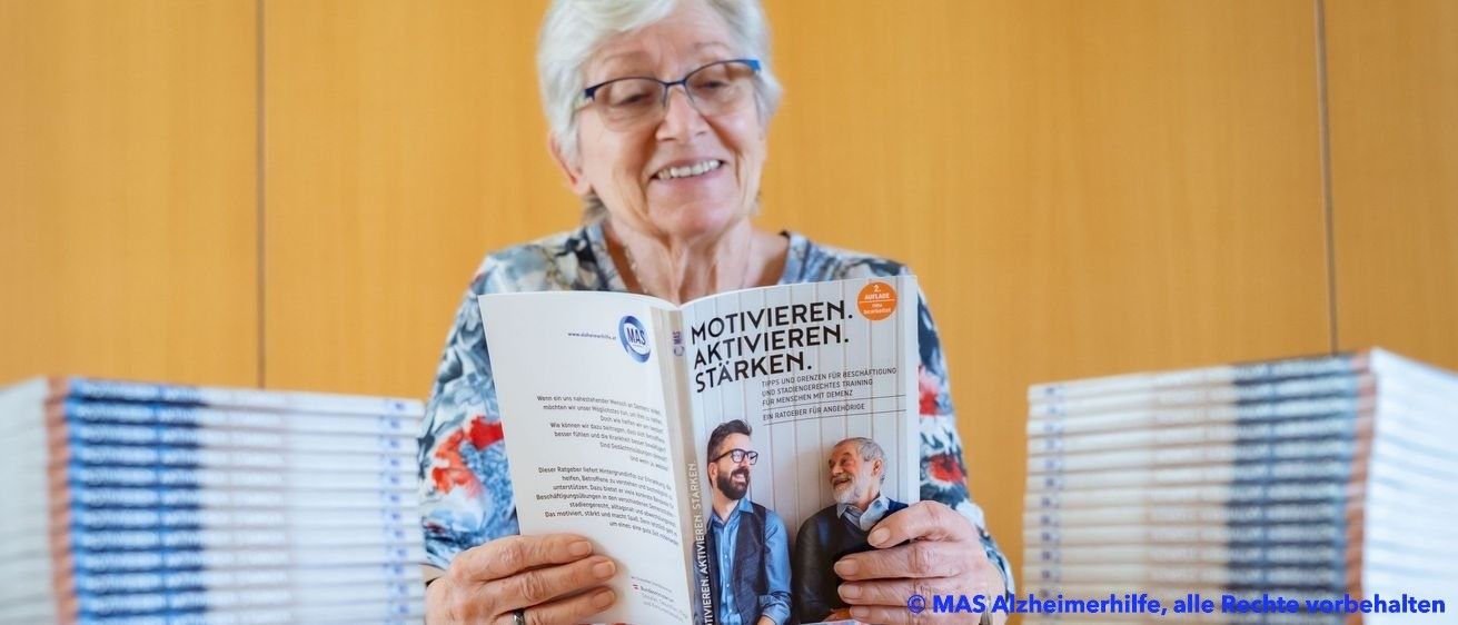 Eine ältere Frau lächelt und liest ein Buch mit dem Titel Motivieren Aktivieren Starken. Auf dem Cover des Buches sind zwei Männer abgebildet. Dahinter befinden sich Stapel desselben Buches.