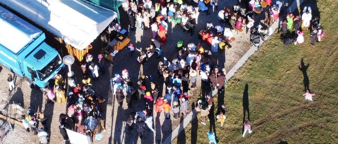 Eine Menschenmenge versammelt sich im Freien, mit einem grünen Zelt in der Mitte. Viele Menschen sitzen an Tischen. Ein blauer Lastwagen ist in der Nähe geparkt. Schatten werfen sich auf den Boden.