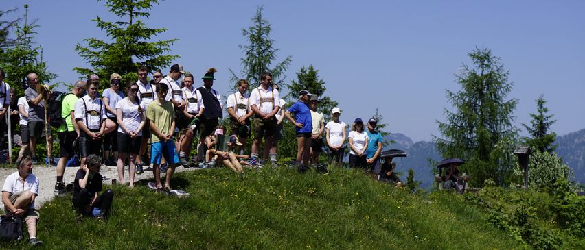Eine Gruppe von Menschen, einige in traditioneller bayerischer Kleidung, steht auf einem grasbewachsenen Hügel unter einem klaren Himmel, möglicherweise bei einem Festival oder einer Veranstaltung. Im Hintergrund befinden sich Bäume und Berge.