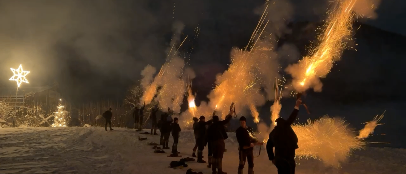 Eine Gruppe von Menschen versammelt sich auf einem verschneiten Hügel in der Nacht und lässt Feuerwerkskörper in den Himmel steigen. Der Boden ist mit Schnee bedeckt und ein Weihnachtsbaum ist in der Ferne zu sehen.
