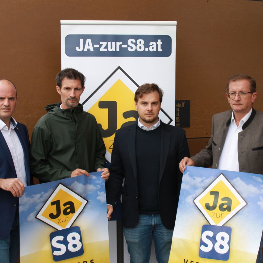 Vier Männer stehen nebeneinander und halten Schilder mit der Aufschrift 'Ja zur S8'. Das Schild im Hintergrund hat ein gelbes Dreieck und steht 'JA-zur-S8.at'.