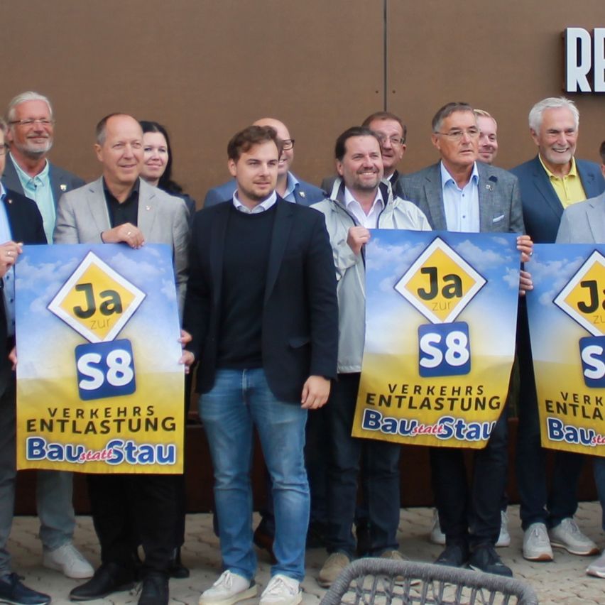 Eine Gruppe von Männern hält Schilder vor einem Gebäude, auf dem RE steht. Sie alle lächeln. Einige von ihnen tragen Brillen und einige tragen Krawatten. Sie tragen alle formelle Kleidung. Die Schilder, die sie halten, haben die Worte Ja S8 Verkehrsentlastung Bau statt Stau.