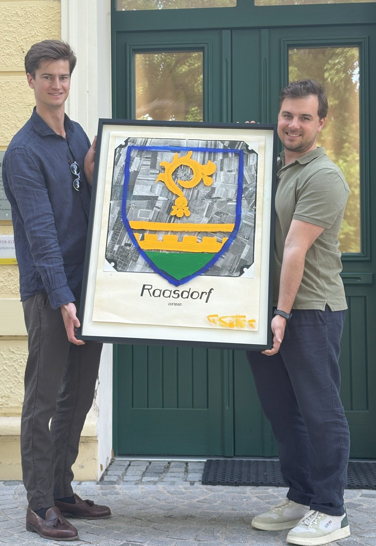 Zwei Männer halten ein gerahmtes Kunstwerk, das das Wappen von Roosdorf darstellt. Sie stehen vor einem Gebäude.