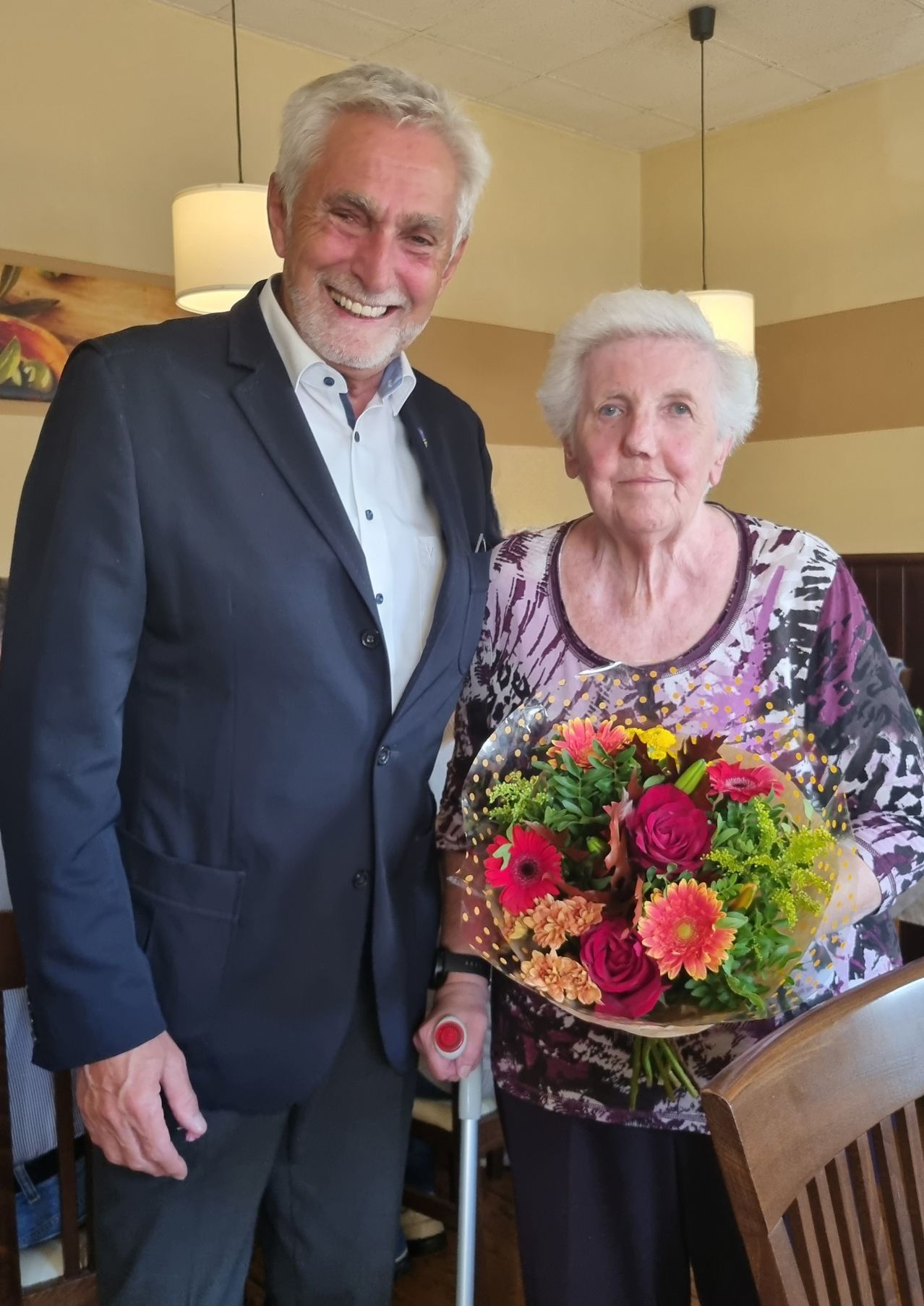 Eine ältere Frau hält einen Blumenstrauß und steht neben einem lächelnden Mann. Sie sind in einem Restaurant mit Holzstühlen und einer gelben Wand.