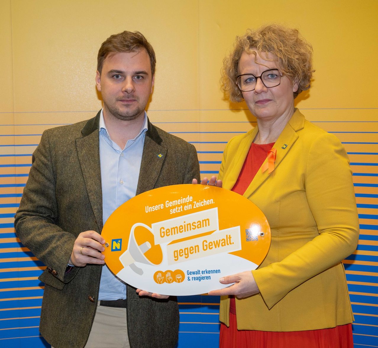 Ein Mann und eine Frau halten ein Schild mit der Aufschrift 'Gemeinsam gegen Gewalt.' Sie stehen vor einem gelben Hintergrund mit blauen Streifen.