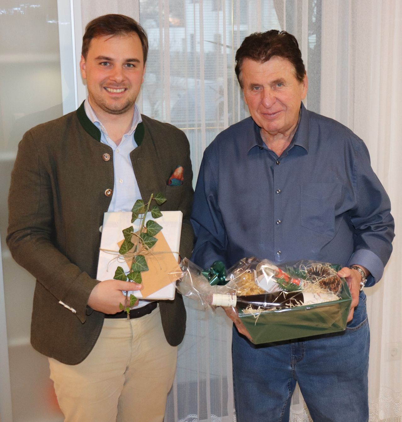 Zwei Männer stehen lächelnd vor einem weißen Vorhang. Ein Mann hält ein eingewickeltes Geschenk mit grünen Blättern und der andere hält eine Box mit einer Weinflasche und anderen Gegenständen.