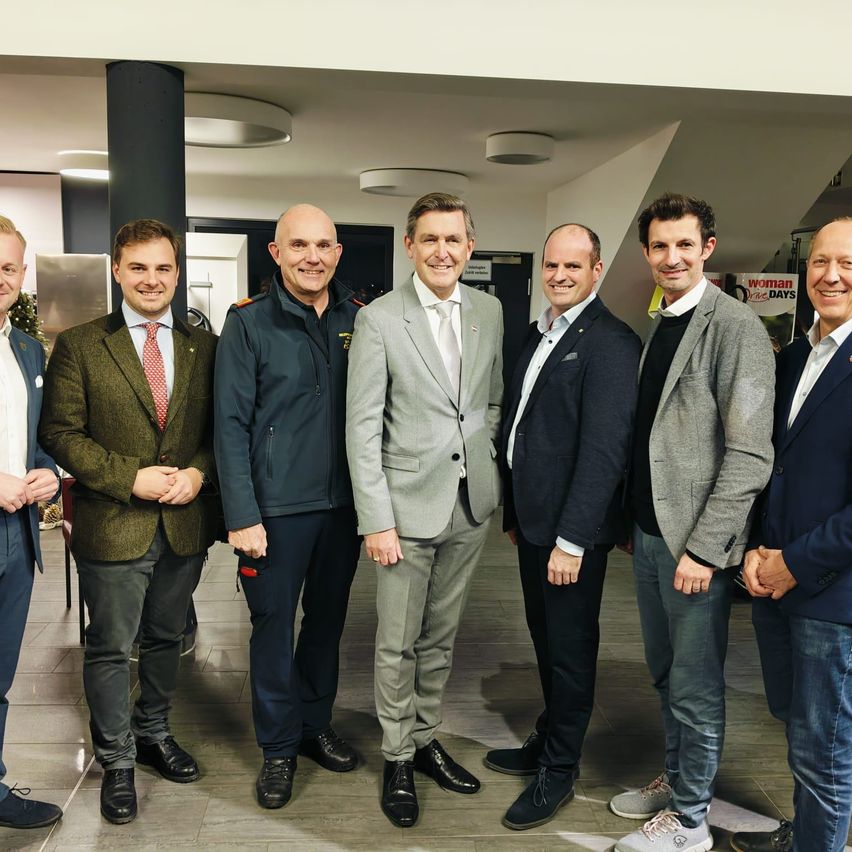 Sieben Männer in Anzügen und Krawatten stehen in einer professionellen Umgebung zusammen und lächeln für ein Foto. Zwei Männer in der Mitte tragen graue Anzüge.