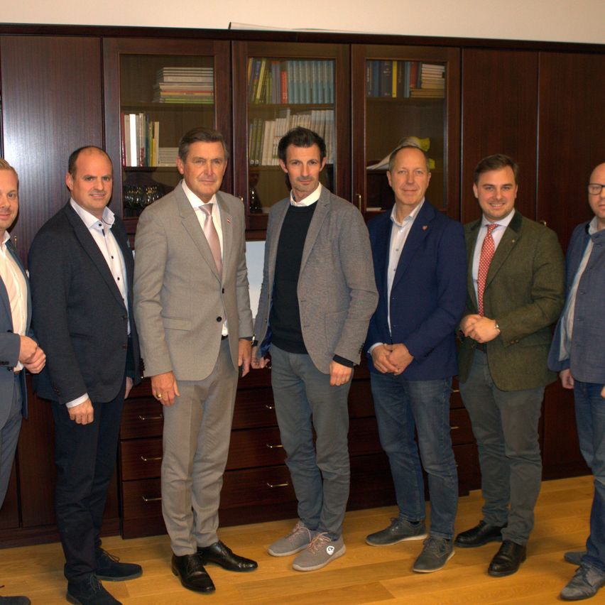 Sieben Männer in Anzügen stehen in einer Reihe in einer professionellen Umgebung. Sie lächeln und schauen in die Kamera. Dahinter steht ein Holzschrank mit Regalen und Büchern.