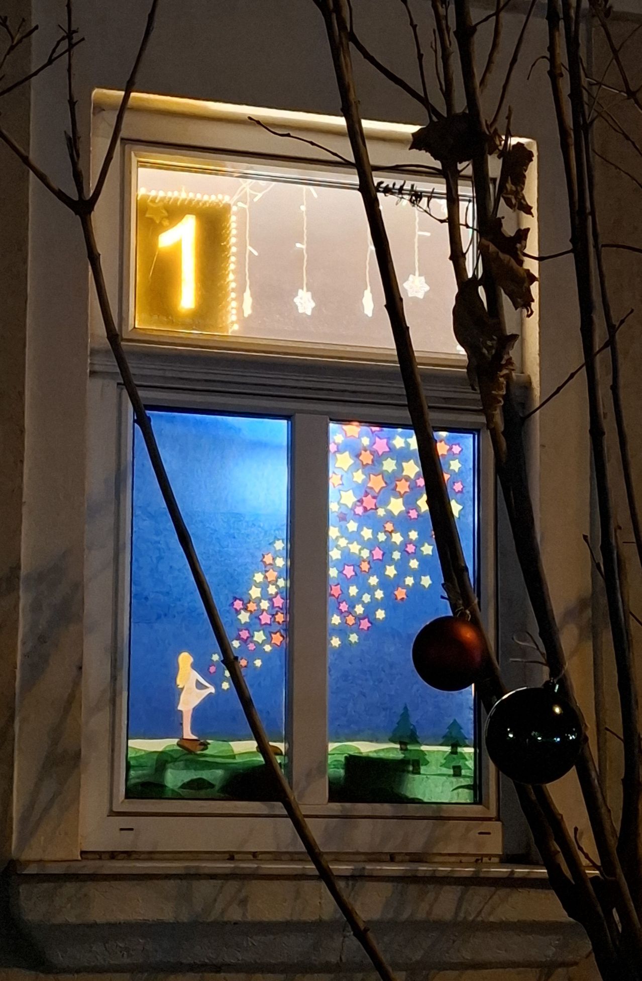 Ein Fenster zeigt eine festliche Szene mit einer Figur, die Sterne pustet, und einem Weihnachtsbaum, mit Dekorationen darüber und Ornamenten darunter.