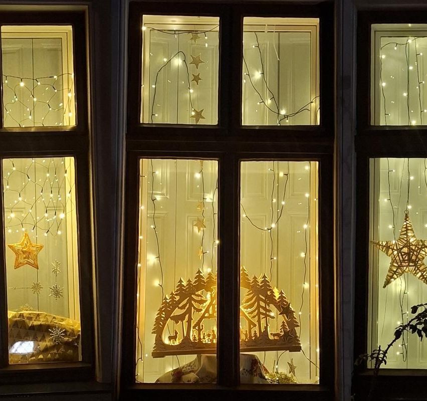 Ein Fensterdisplay mit Lichterketten und Sternen, das einen Weihnachtsbaum und eine Krippe zeigt. Die Szene ist mit warmen gelben Lichtern beleuchtet.