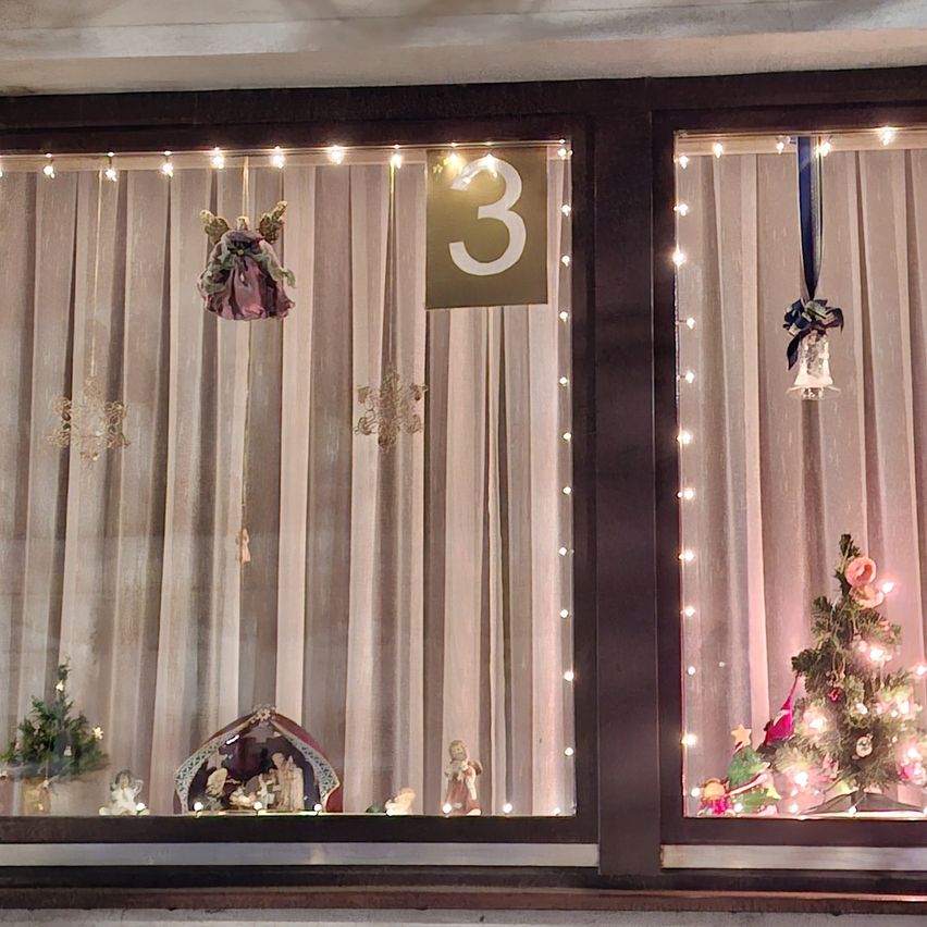 Eine Fensterdekoration mit Weihnachtsschmuck, darunter eine Krippe, Engelfiguren und ein Weihnachtsbaum, alles beleuchtet von Lichterketten. Das Fenster ist in zwei Bereiche unterteilt, jeder mit eigenen einzigartigen Dekorationen.