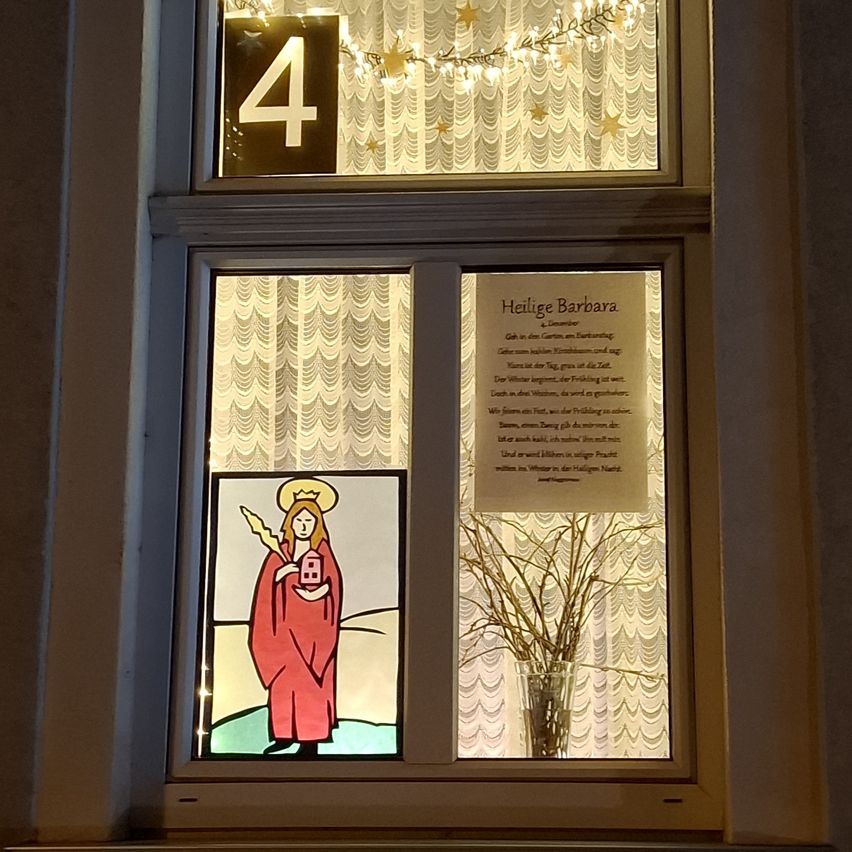 Ein Fenster zeigt ein Buntglas von der Heiligen Barbara, die ein Haus hält, mit 'Heilige Barbara' auf einem weißen Plakat. Oben leuchtet ein '4'-Schild mit goldenen Sternen. Ein Vasen mit kahlen Zweigen ist ebenfalls sichtbar.