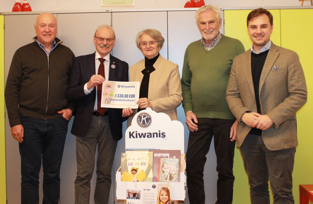 Fünf Personen posieren für ein Foto und halten ein Plakat mit dem Text 1.530,00 EUR und einem Kiwani International Logo. Sie stehen vor einem Poster und einem Bücherregal.