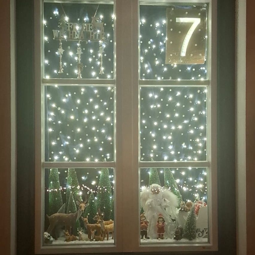 Eine Weihnachtsdekoration an einem Fenster mit der Nummer 7, mit einem weißen Engel, Rehen und einem Schlitten mit dem Weihnachtsmann und schneebedeckten Kiefern.