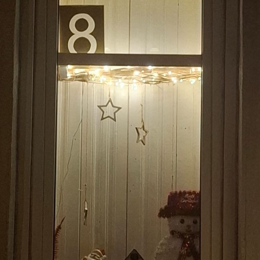 Ein Fensterdisplay mit der Nummer 8 darüber. Das Fenster ist mit einer Lichterkette, einem Stern und einem Schneemann dekoriert. Der Schneemann hat einen Hut und eine Krawatte.