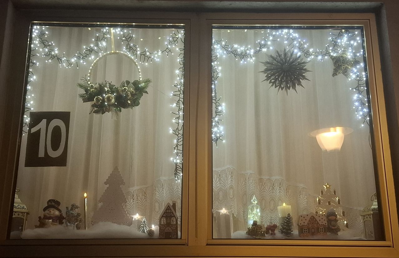 Ein Fensterdisplay mit weißen Vorhängen und einem Kranz. Der Fensterrahmen ist mit Lichterketten und Weihnachtsdekorationen geschmückt, darunter eine Kerze, ein Lebkuchenhaus und ein Weihnachtsbaum. Das rechte Fenster hat zusätzliche Ornamente.
