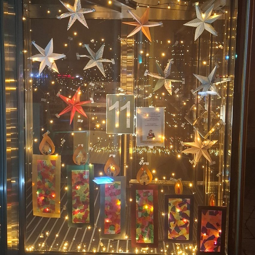 Eine Glasvitrine mit beleuchteten Kerzen und Papiersternen. Die Kerzen haben bunte Muster, und es gibt hängende Sterne und Lichter um die Ausstellung. Eine Nummer 11 ist prominent angebracht.