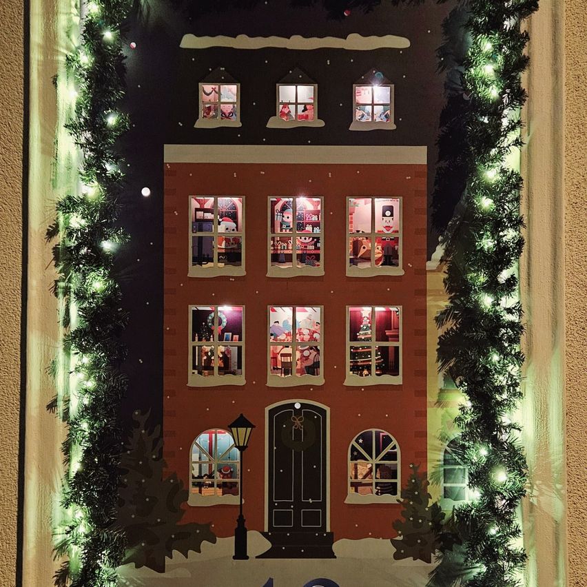 Ein dekoratives Weihnachtshaus mit beleuchteten Fenstern und einer Girlande, geschmückt mit Lichtern und Grün. Jede Etage hat eine einzigartige Weihnachtsszene, die durch die Fenster sichtbar ist.