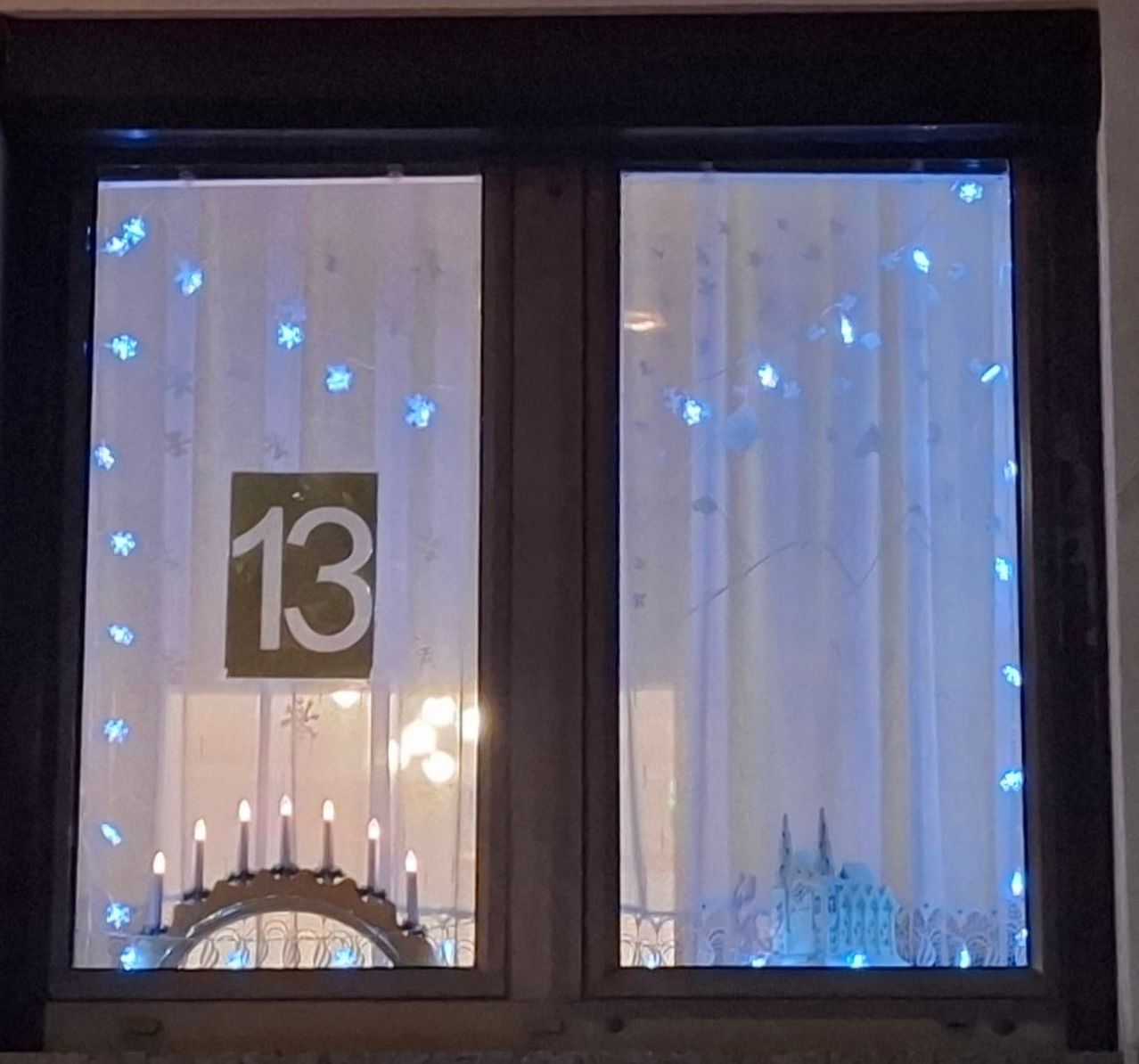 Ein Fenster mit der Nummer 13. Schneeflocken und blaue Lichter sind am Fenster. Ein Kerzenständer mit Kerzen und blauen Lichtern steht vor dem Fenster.