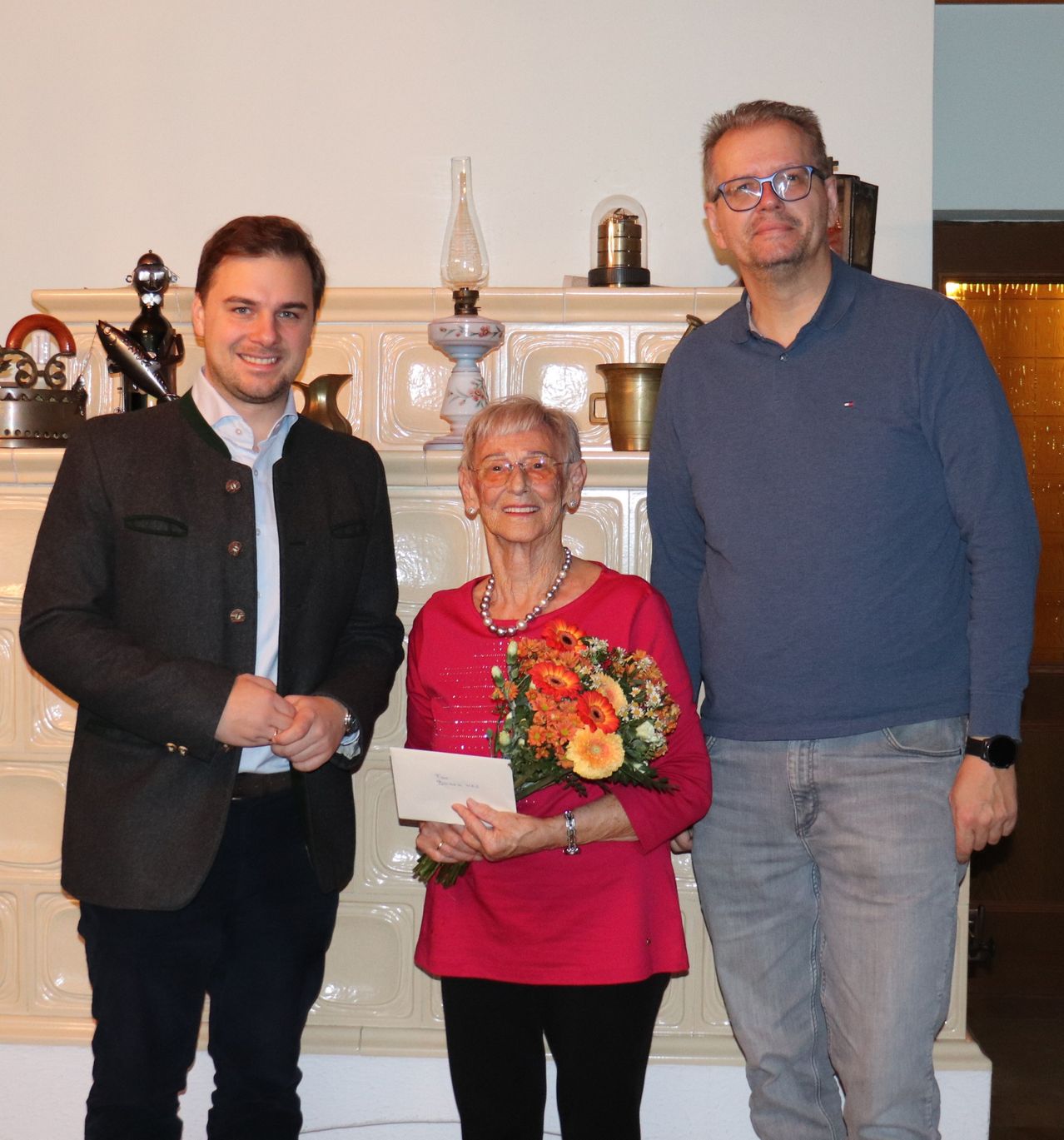 Drei Personen stehen zusammen vor einer gefliesten Wand, wobei eine ältere Frau einen Blumenstrauß hält und zwei Männer auf beiden Seiten von ihr stehen.