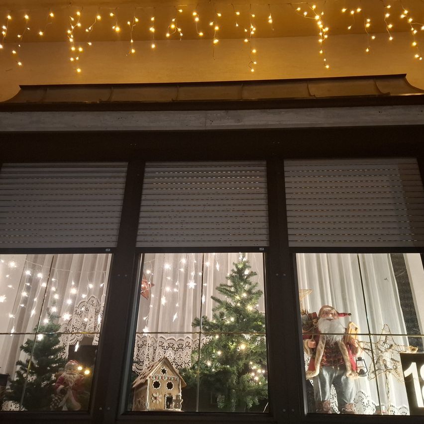 Das Bild zeigt ein festlich geschmücktes Fenster mit einer Weihnachtsmannfigur, einem Lebkuchenhaus und einem Weihnachtsbaum, beleuchtet von Lichterketten.