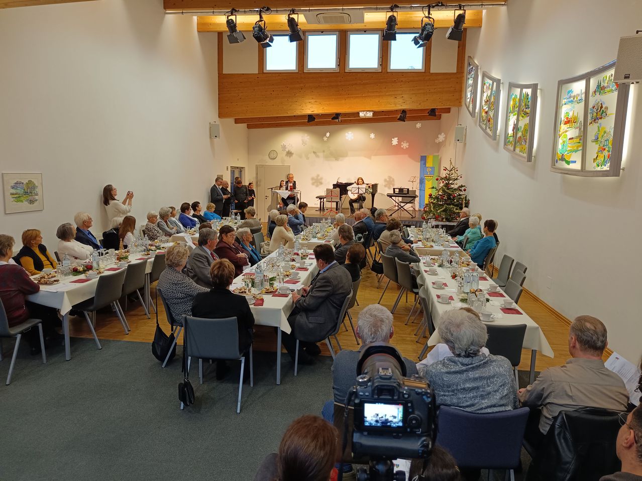 Ein Saal beherbergt ein Essereignis mit einem Weihnachtsbaum und einem Musiker. Viele Menschen sitzen um Tische herum, einige stehen und ein Fotograf hält den Moment fest.