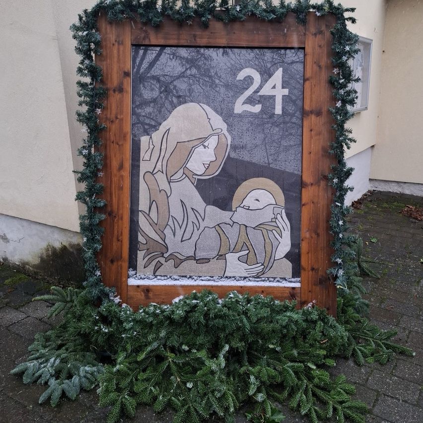 Ein hölzern gerahmtes Bild mit der Nummer 24 und einem Bild einer Frau, die ein Baby hält, liegt neben einem Gebäude auf dem Boden.