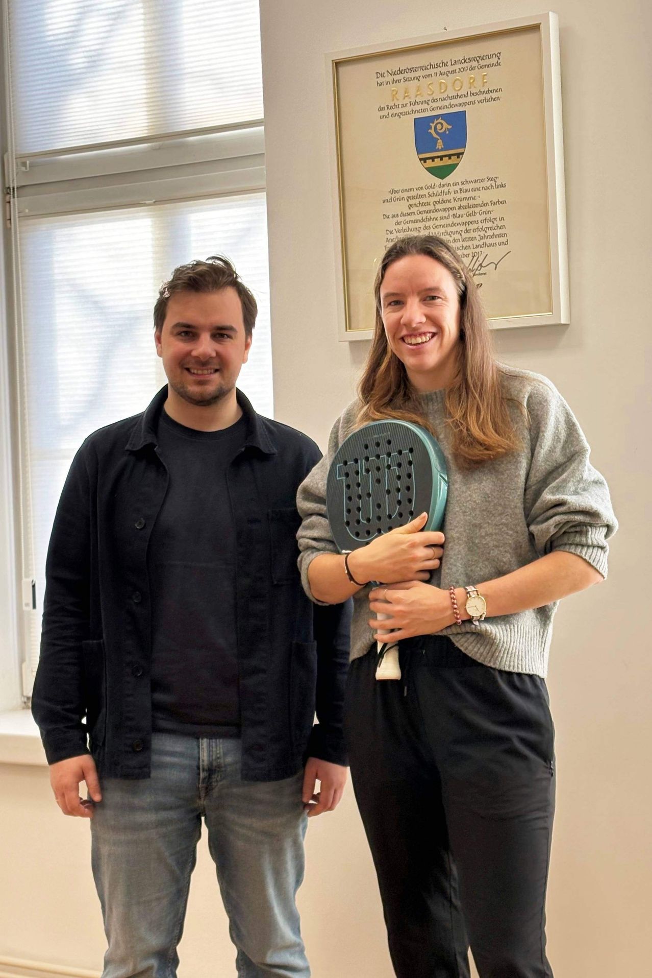 Ein Mann und eine Frau stehen drinnen. Die Frau hält einen Tennisschläger und lächelt. Hinter ihnen ist ein gerahmtes Zertifikat an der Wand.