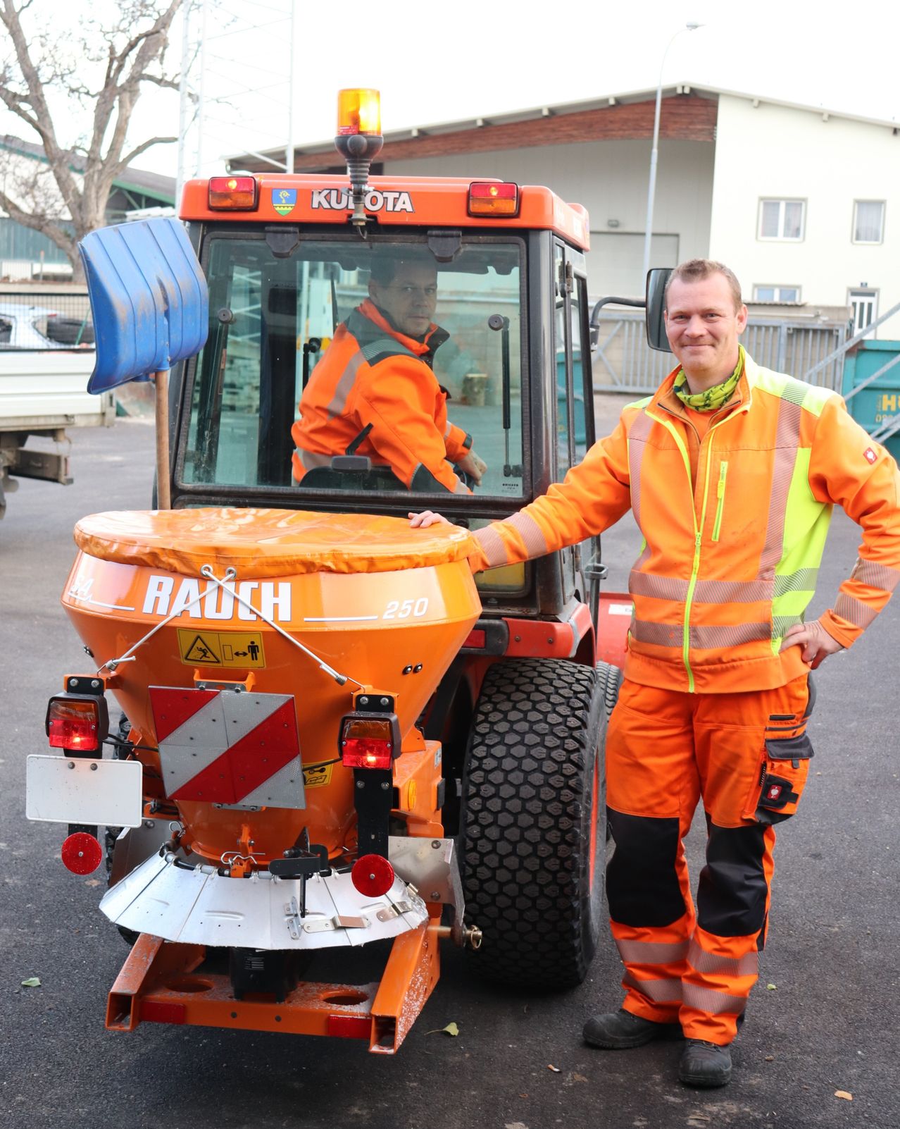 Zwei Männer in orangefarbenen Sicherheitsanzügen stehen neben einem Kubota-Traktor. Einer sitzt im Inneren des Traktors und der andere steht neben der Tür und lächelt.