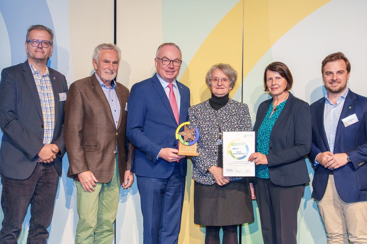 Vier Personen stehen gemeinsam, zwei Männer und zwei Frauen, präsentieren ein Zertifikat und einen Preis. Sie sind formell gekleidet.