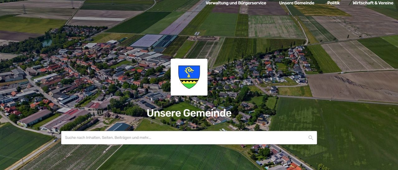 An aerial view of a rural community with houses and buildings, surrounded by green fields and trees, with a watermark that reads Unsere Gemeinde.