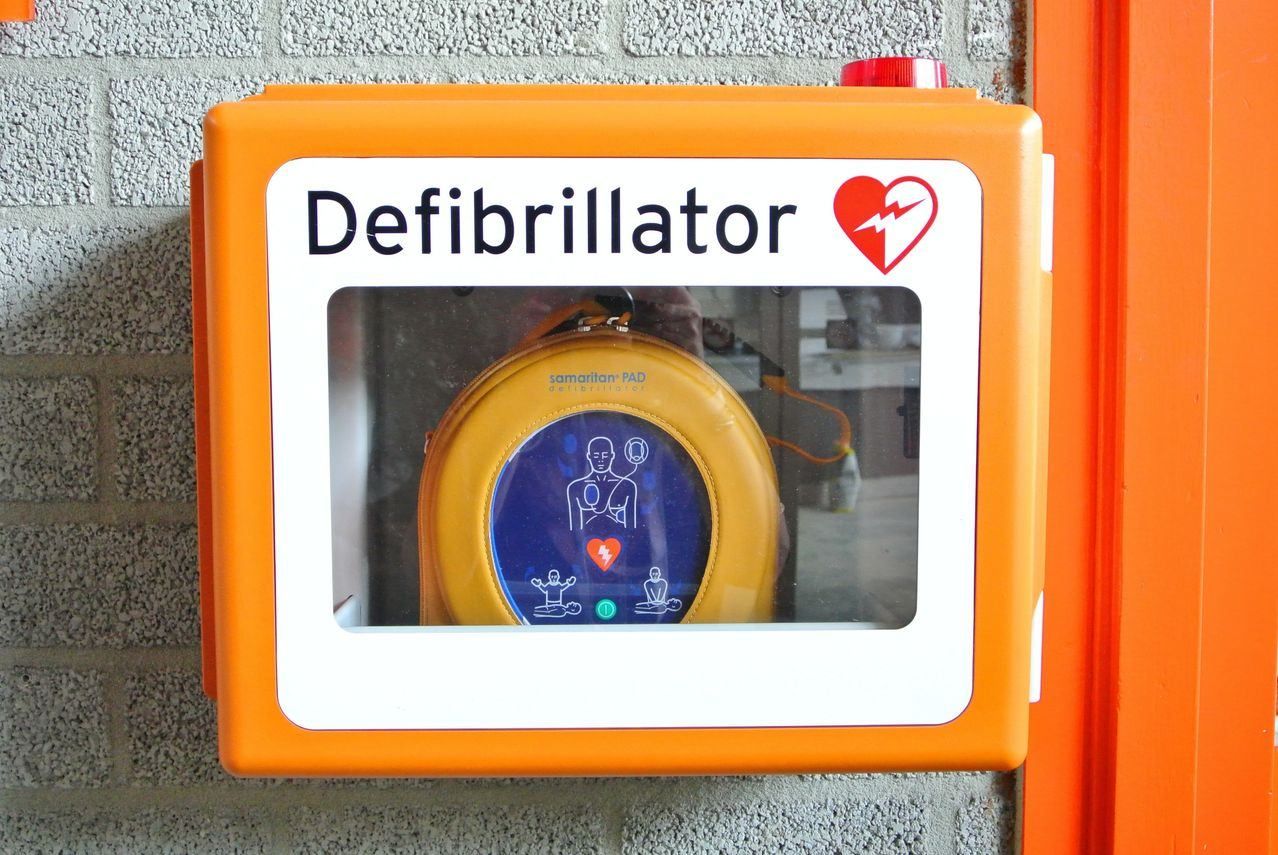 Orangefarbene Defibrillator-Box an der Wand montiert mit einem Samaritan PAD im Inneren. Das Gerät zeigt Symbole von Menschen und einem Herzen mit Blitz.