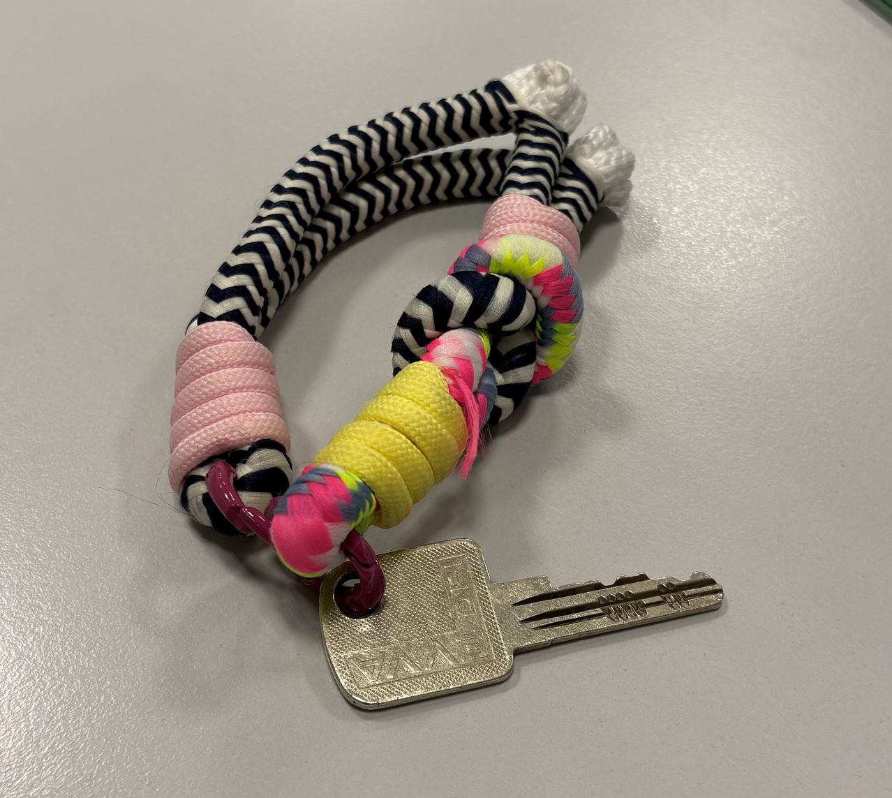 Ein farbenfroher, handgefertigter Schlüsselanhänger mit Zickzackmustern in rosa, gelb und schwarz. Ein silberner Schlüssel mit Text ist befestigt.