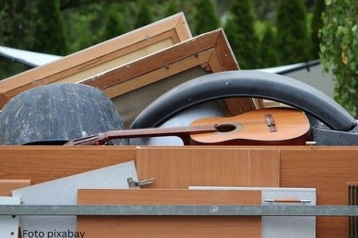 Ein Haufen ausrangierter Gegenstände, darunter eine Gitarre, ein Reifen und ein Holzstuhl, wird auf einen Lastwagen geladen. Im Hintergrund sind grüne Bäume und ein klarer Himmel zu sehen.