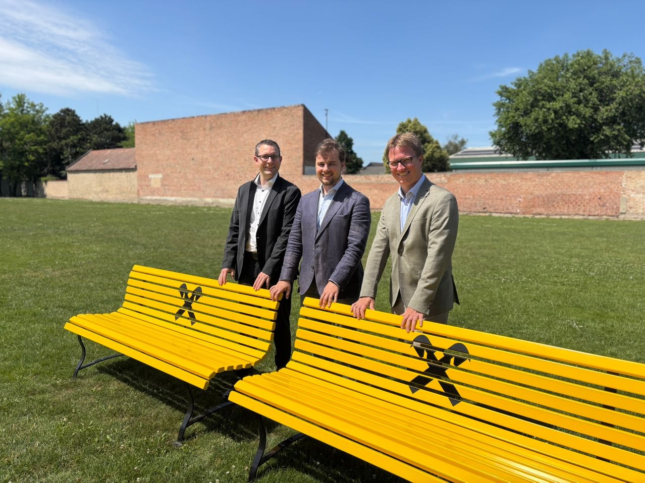 Three men stand next to two bright yellow benches in a grassy field with a brick building in the background.